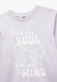 Lavender sweatshirt met witte bloemgrafieken en tekst. De tekst luidt "Voed je ZIEL" en "Voed je GEEST" in verschillende lettertypes. Zacht, glad materiaal.
