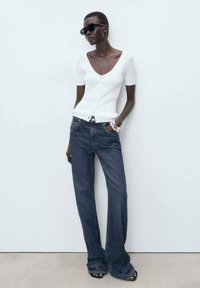 White fitted top with short sleeves, V-neck, paired with dark blue wide-leg jeans, and black sunglasses. Smooth texture and casual design.