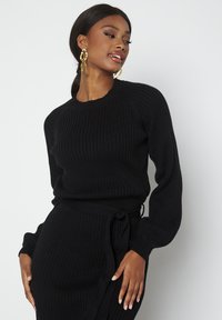 Maglione nero a coste con maniche lunghe e leggermente a sbuffo, dal design aderente. Accentuato da un fiocco coordinato in vita.