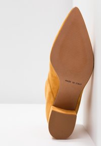 Chaussure jaune en daim à bout pointu avec semelle texturée marron et talon carré, étiquetée "Fabriquée en Italie", inclinée contre une surface blanche.