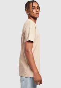 Beige cotton t-shirt with short sleeves, crew neck, and a relaxed fit. Worn with light blue jeans, model facing slightly to the side.