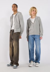 Grå hettegensere med glidelås og frontlommer, kombinert med løstsittende mørke og lyse denimjeans. Sko med flerfarget design gir kontrast.
