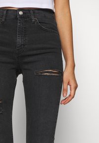 Svart högmidjade jeans med slitna knäbitar, med knäppning fram, femfickorsdesign och subtila blekningar. Slät denimtextur.