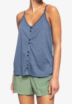 Kvinde iført blå, knaplukket ærmeløs top og lysegrønne shorts, stående mod en ensfarvet hvid baggrund.