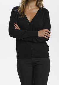 Svart V-hals cardigan med långa ärmar, knäppning och mjuk textur. Kombinerad med svarta byxor för en sammanhängande look.
