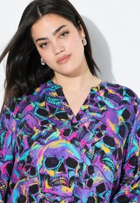 Farbige Bluse mit einem lebhaften Totenkopf-Muster in lila, schwarz und Neonfarben; leichtes Material mit V-Ausschnitt.