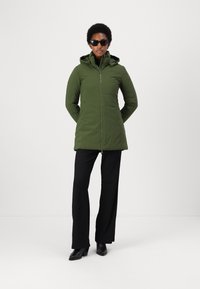 Chaqueta aislante verde con cierre frontal, capucha ajustable, mangas largas y bolsillos laterales, combinada con pantalones negros y zapatos de tacón.