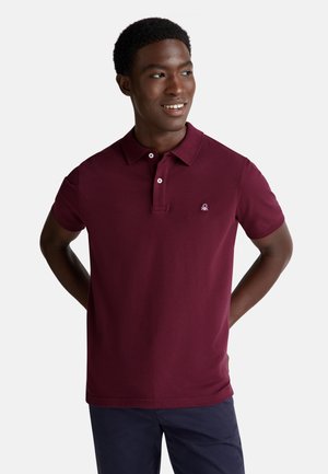 Hombre vestido con una camiseta tipo polo de manga corta color burdeos con botones, pantalones azul marino, de pie con las manos detrás de la espalda, sonriendo y mirando hacia la derecha.