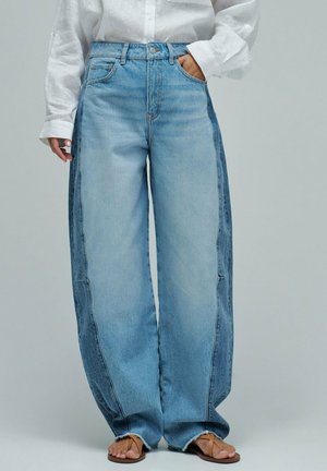 Persona con vaqueros holgados de mezclilla azul de dos tonos con dobladillos deshilachados, camisa blanca de manga larga y sandalias marrones, de pie con una mano en el bolsillo.