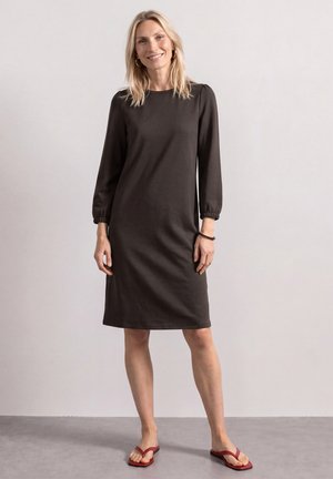 Femme blonde souriante, portant une robe noire à manches longues jusqu'aux genoux et des tongs rouges, debout devant un fond clair uni.
