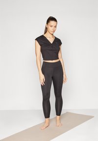 Svart cropped treningsgenser med korte ermer, med tvistdetalj, sammen med høytlivede svarte tights, stående på en lys matte.