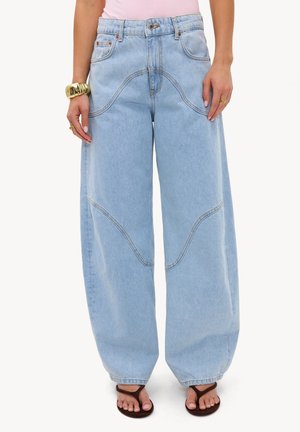 Jeans bleu clair à jambes larges avec des détails de coutures courbes, portés avec des tongs marron et des bracelets dorés, montrés de la taille aux pieds.
