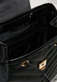 Musta käsilaukku, jossa on tikattu pinta, kultaiset yksityiskohdat ja useita sisätaskuja, mukaan lukien vetoketjullinen osio ja sileä sisävuori.