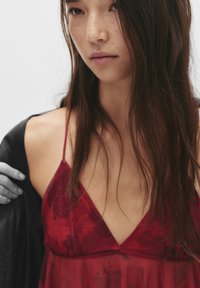 Donna con lunghi capelli scuri che indossa un vestito floreale rosso con spalline sottili, mentre si toglie un giubbotto nero dalla spalla su uno sfondo semplice.