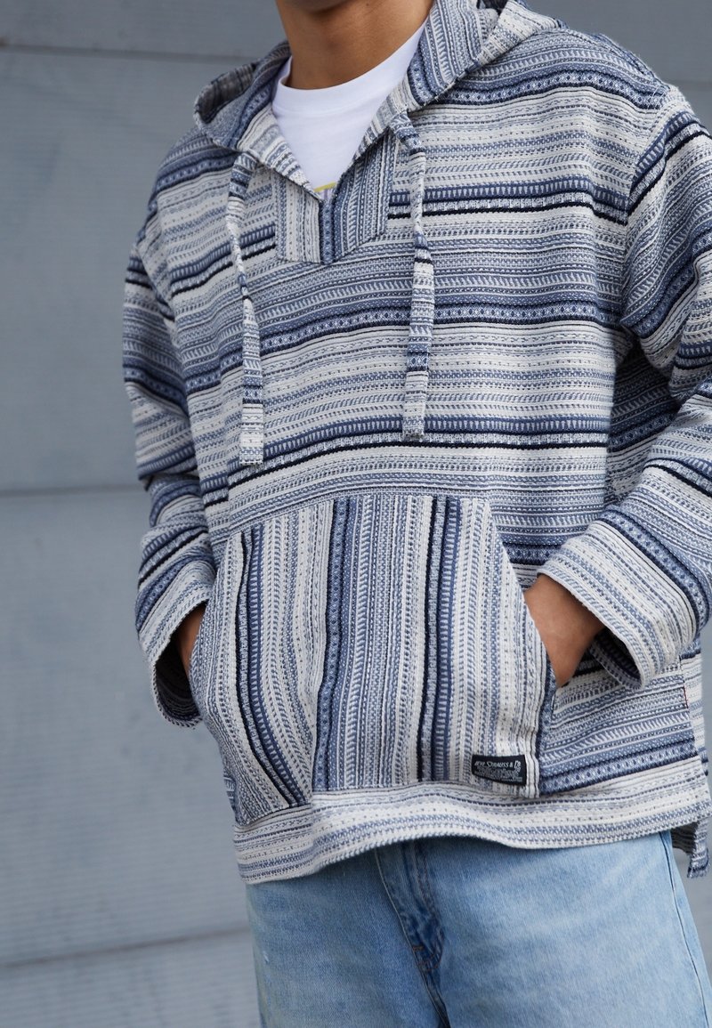 Personne portant un sweat à capuche rayé bleu et blanc avec une poche avant et des cordons, associé à un jean bleu clair. Mains dans les poches.
