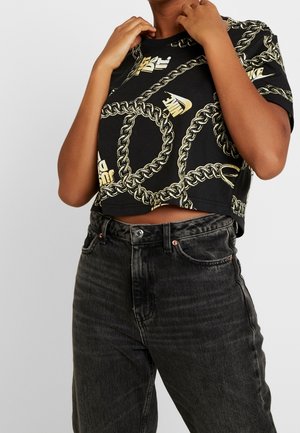 Person wearing a black cropped T-shirt with gold chain and "Nike" logos, paired with high-waisted faded black jeans.