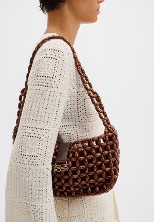 Άτομο που φοράει κρεμ πλεκτό πουλόβερ με τετράγωνα σχέδια, κρατώντας μια καφέ τσάντα από ξύλινες χάντρες με δερμάτινο κούμπωμα στον ώμο.