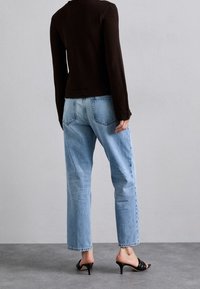 Schwarzes gestricktes Langarmshirt, hellblaue gerade Jeans und schwarze Absatzsandalen. Bemerkenswerte Details sind der strukturierte Stoff und die umgeschlagenen Säume.