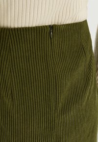 Jupe en velours côtelé vert olive avec une texture côtelée verticale. Dispose d'une fermeture éclair discrète à la taille, mettant en valeur un design ajusté.