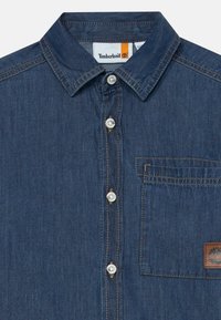 Denimskjorte med spiss krage, sølvknapper, brystlomme, kontraststikking og Timberland-logo. Mørk blå farge.