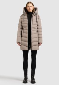 Lichtbeige pufferjas met een capuchon, voorzien van horizontale stiknaden, ritsluiting en trekkoordaccenten. G gedragen over een zwarte outfit.