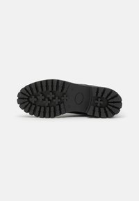 Semelle de chaussure noire avec un motif de crampons profond, comprenant des crampons rectangulaires et en forme de croix pour une meilleure adhérence et traction.