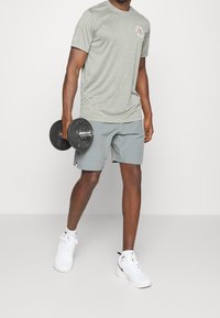 Šedé sportovní tričko s logem, doplněné světle šedými šortkami. Model drží černou činku a má na sobě bílé tenisky. Jednoduchý, funkční design.