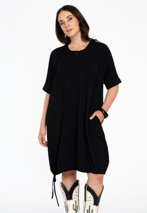 Schwarz, lockeres Kleid mit kurzen Ärmeln, Knopfdetails, Taschen und einem gerafften Saum. Kombiniert mit weißen Cowboy-Stiefeln mit Kaktus-Design.