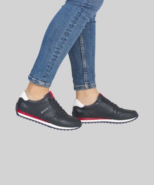 Personne portant des baskets noires avec des accents rouges et blancs, associées à un jean skinny bleu retroussé, debout sur un fond gris clair.