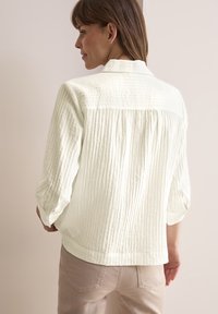 Camicia button-up di un crema chiaro con un tessuto texturizzato, maniche arrotolate e un design con pieghe sul retro. Abbinata a pantaloni beige chiaro.