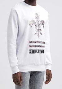 Bílá mikina s kulatým výstřihem, zdobená květinovým motivem fleur-de-lis a černým textem "CXMINNL DIVMGE" nad dvěma vodorovnými pruhy.