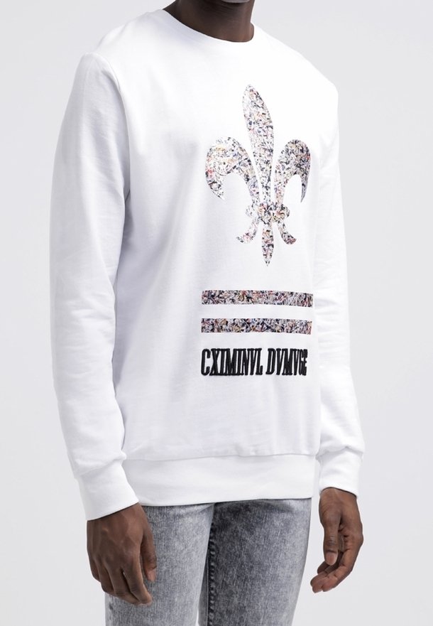 Bílá mikina s kulatým výstřihem, zdobená květinovým motivem fleur-de-lis a černým textem "CXMINNL DIVMGE" nad dvěma vodorovnými pruhy.