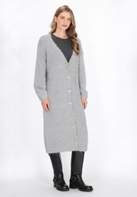 Grauer, flauschiger Cardigan mit V-Ausschnitt, langen Ärmeln und goldenen Knöpfen auf der Vorderseite, kombiniert mit einem dunklen Oberteil und knielangen schwarzen Stiefeln.