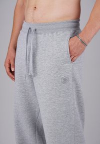 Man wearing light grey sweatpants with drawstring, hand in right pocket, and silver chain bracelet on right wrist, against plain background.