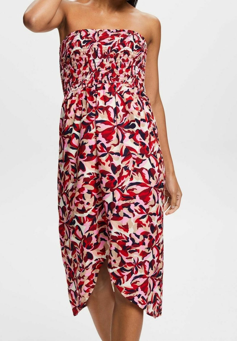 Femme portant une robe sans bretelles, longueur genou, avec un motif floral rouge, rose et noir et un corsage froncé, debout devant un fond uni.