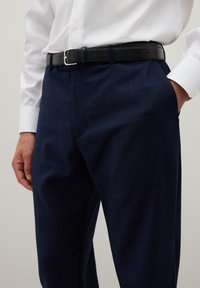 Pantalones de cuadros en tonos marinos, confeccionados en una tela de textura suave, con un cinturón de cuero negro y hebilla plateada. Camisa blanca con cuello visible.