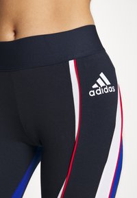 Navy leggings valmistettu joustavasta materiaalista, joissa on valkoiset, punaiset ja siniset sivusuunnat, sekä Adidas-logo vasemmalla reidessä.