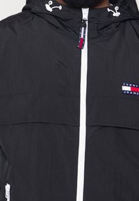 Czarna lekka kurtka z białym zamkiem błyskawicznym, z kapturem i naszywką z logo na piersi. Gładka faktura o sportowym designie.