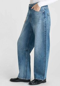 Personne portant un jean large bleu clair et des mocassins noirs, la main dans la poche, se tenant contre un fond blanc.