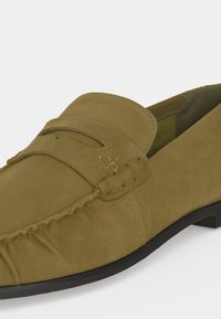 Mocassin en daim beige avec des détails de couture, une petite bride sur le dessus, et un logo doré "SM" sur le côté extérieur.