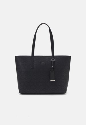 Sac cabas noir Calvin Klein avec motif monogramme, deux bandoulières et une étiquette suspendue sur un fond blanc.