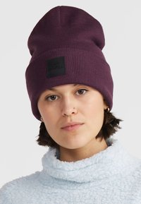 Violetti ribattu pipo, jossa on musta logo-etiketti, pehmeä rakenne ja rento muotoilu. Päällä vaalean sininen fleecestä valmistettu korkea kaulaosa.