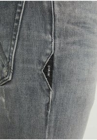 Grå denimjeans med en närbild av sido sömmen med en svart etikett som läser "CHAIM." Anmärkningsvärd textur med lätt slitning och slät yta.