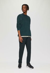 Donkergroen ribbeltrui met ronde hals, gecombineerd met donkergroene tapered broek en zwarte sportschoenen met lichte accenten.