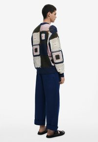 Strickpullover mit einem mehrfarbigen Patchwork-Design aus großen Quadraten, kombiniert mit lässigen dunkelblauen Hosen und flachen schwarzen Sandalen.