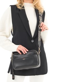 Kvinden har en hvid sweater og en sort vest på, og hun holder en sort læder crossbody taske med en leopardmønstret rem.