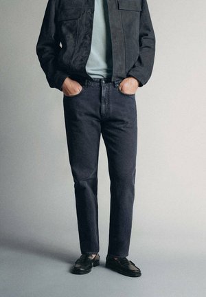 Personne portant un jean noir, une veste noire, une chemise claire et des mocassins noirs, debout les mains dans les poches devant un fond uni.