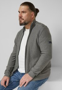 Grauer Cardigan mit Reißverschluss, mit geripptem Kragen und Bündchen, aus weichem Stoff, getragen über einem weißen T-Shirt, kombiniert mit blauen Jeans.