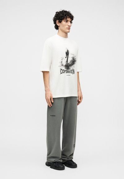 Άνδρας με σγουρά μαλλιά που φοράει άσπρο υπερμεγέθες μπλουζάκι με γραφικό κύκνου και την επιγραφή «Κοπεγχάγη», γκρι φόρμα και μαύρα αθλητικά παπούτσια.