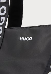 Melns auduma soma ar drosmīgu, logo reljefā izstrādāta siksna un spīdīgu "HUGO" etiķeti baltā krāsā priekšpusē. Gluda tekstūra, minimālisma dizains.